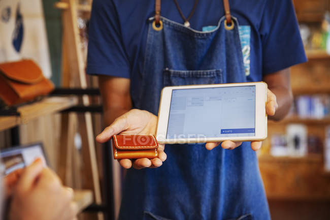 Man wearing blue apron standing in a leather shop, holding leather purse and digital tablet. — Stock Photo