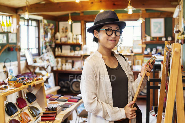 Японська жінка в капелюсі й окулярах стоїть у шкіряній крамниці, тримаючи шкіряний пояс.. — стокове фото