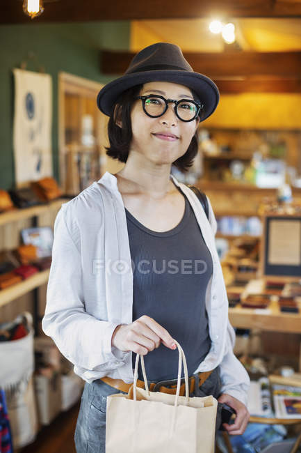 Mulher japonesa vestindo chapéu e óculos de pé em uma loja de couro, segurando saco de compras, sorrindo na câmera . — Fotografia de Stock