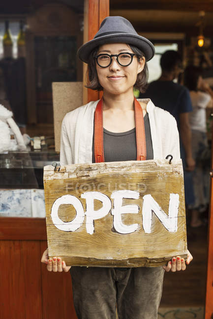 Femme japonaise portant chapeau et lunettes debout devant une boutique de cuir, tenant signe ouvert . — Photo de stock