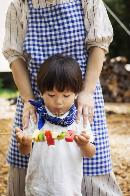 Japanese woman and boy standing outdoors, holding skewer with fresh kiwi and melon. — Stock Photo