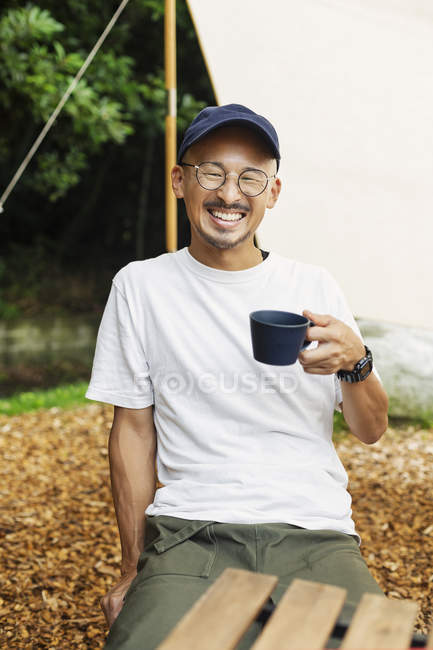 Lächelnder Japaner mit Baseballkappe und Brille sitzt draußen an einem Tisch und trinkt eine Tasse Kaffee. — Stockfoto