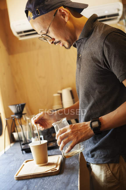 Japaner mit Baseballkappe und Brille steht in einem Öko-Café und bereitet eine Tasse Kaffee zu. — Stockfoto