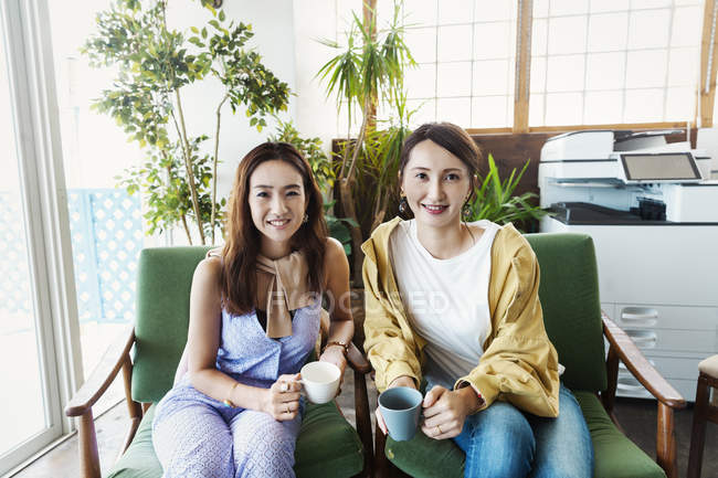 Two female Japanese professionals with cups of coffee sitting in a co-working space in chairs, smiling in camera. — Stock Photo