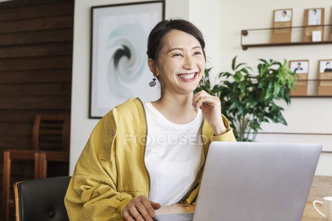 Mulher japonesa sorridente sentada a uma mesa em um espaço de co-trabalho, usando computador portátil . — Fotografia de Stock