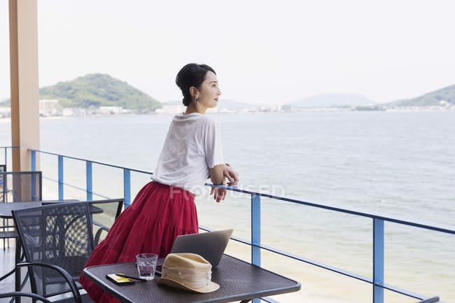 Japanerin steht auf Balkon eines Co-Working Space, Laptop auf Tisch. — Stockfoto
