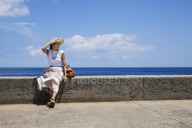 Japanerin mit Hut sitzt auf einer Mauer am Meer. — Stockfoto
