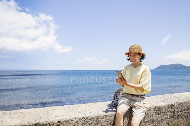 Mulher japonesa usando chapéu sentado em uma parede por oceano . — Fotografia de Stock