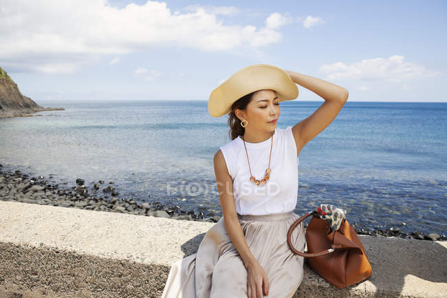 Japonesa mujer usando sombrero sentado en una pared por océano
. - foto de stock