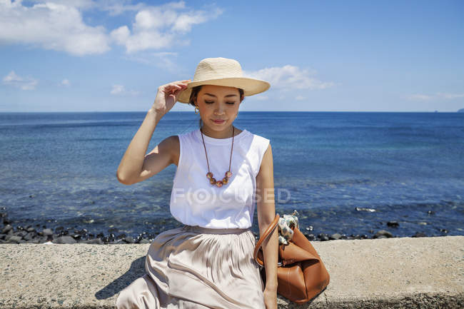 Mulher japonesa usando chapéu sentado em uma parede por oceano . — Fotografia de Stock