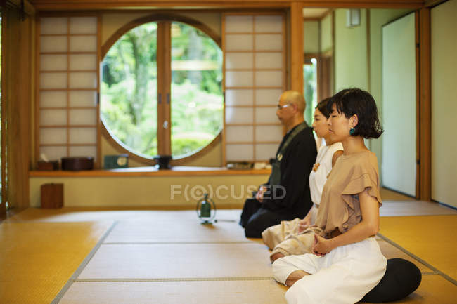 Dos mujeres japonesas y un sacerdote budista arrodillados en el templo budista, rezando . - foto de stock