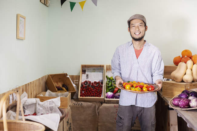 Homem japonês sorrindo vestindo boné de pé na fazenda loja, segurando tigela com pimentas frescas, sorrindo na câmera . — Fotografia de Stock
