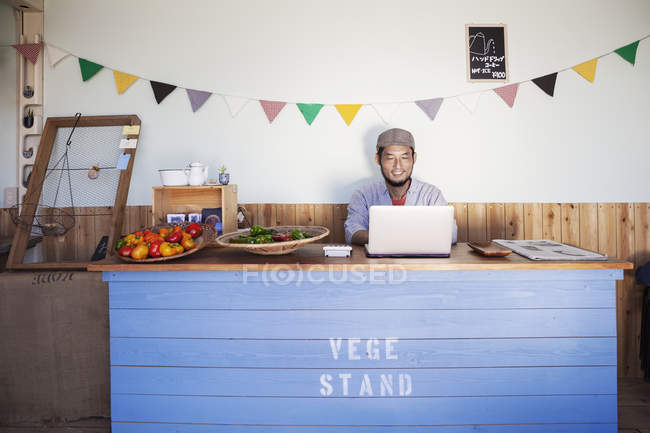 Japanese man sitting behind counter of a farm shop, working on laptop computer. — Stock Photo