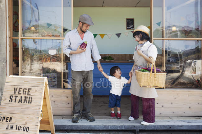 Japonés hombre, mujer y niño de pie fuera de una granja tienda, tomados de la mano . - foto de stock