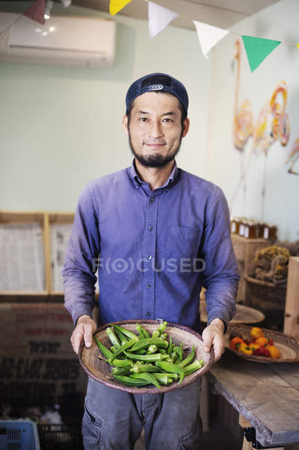 Sonriente hombre japonés con gorra de pie en la tienda de la granja, sosteniendo un tazón con okra fresca . - foto de stock
