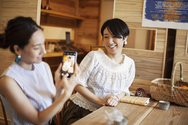 Duas mulheres japonesas sentadas em uma mesa em um café vegetariano, usando telefone celular e tirando fotos . — Fotografia de Stock