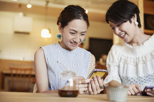 Dos mujeres japonesas sentadas en una mesa en un café vegetariano, usando teléfono móvil . - foto de stock