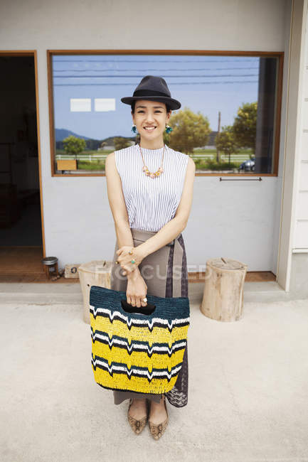 Portrait of smiling Japanese woman wearing hat and holding shopping bag, looking in camera. — Stock Photo