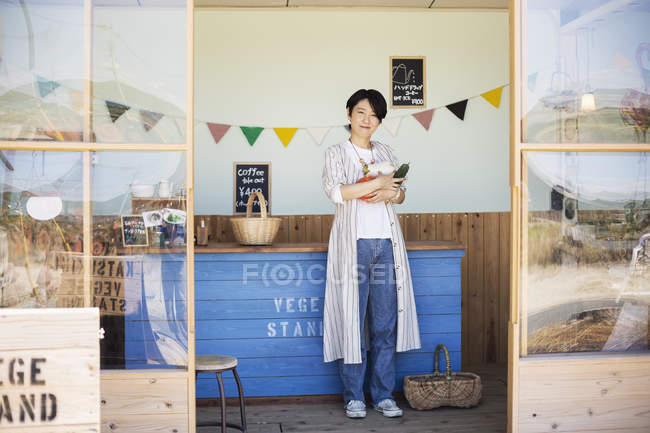 Japanese woman standing in a farm shop, holding vegetables, smiling in camera. — Stock Photo
