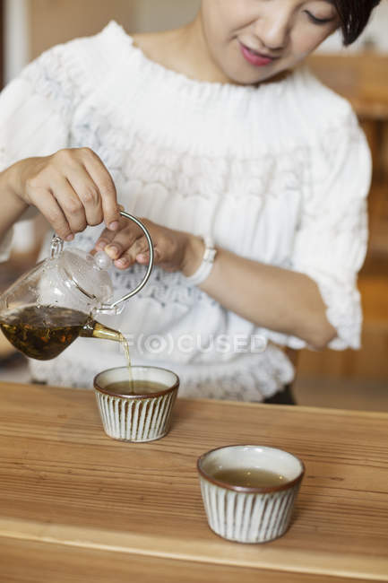 Japanese woman sitting at a table in a vegetarian cafe, pouring tea. — Stock Photo