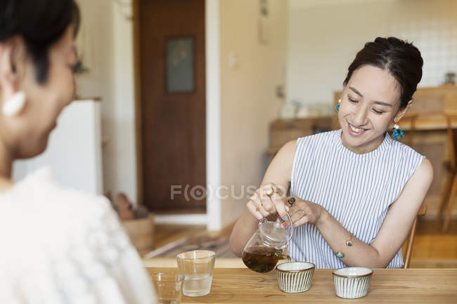 Two Japanese women sitting at a table in a vegetarian cafe, pouring tea. — Stock Photo