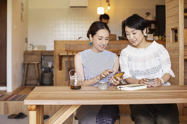 Zwei japanische Frauen sitzen an einem Tisch in einem vegetarischen Café und benutzen ein Mobiltelefon. — Stockfoto