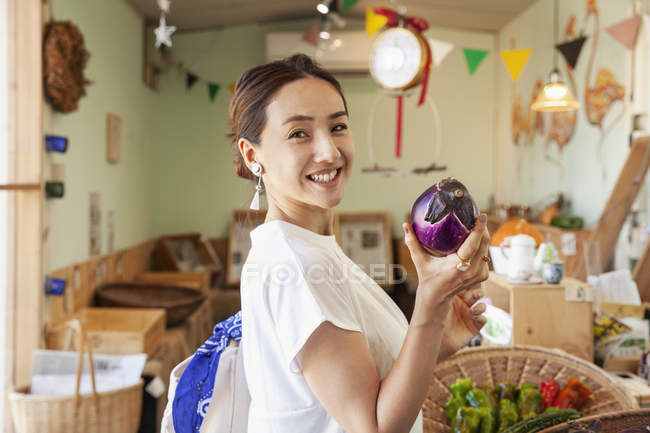 Sonriendo mujer japonesa de pie en una tienda de granja, sosteniendo berenjena, mirando a la cámara . - foto de stock