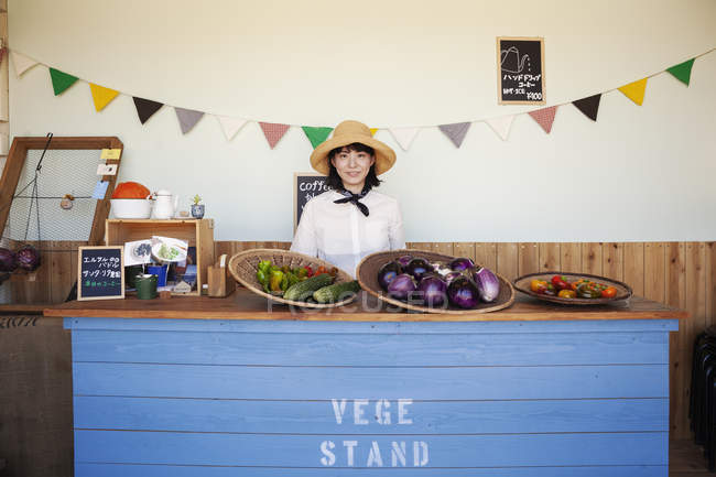 Japanese woman in hat standing behind counter in a farm shop, smiling in camera. — Stock Photo