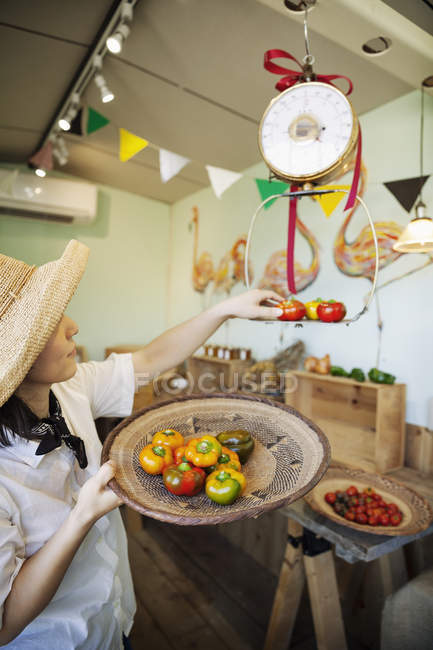 Japanese woman in hat working in a farm shop, weighing fresh peppers. — Stock Photo