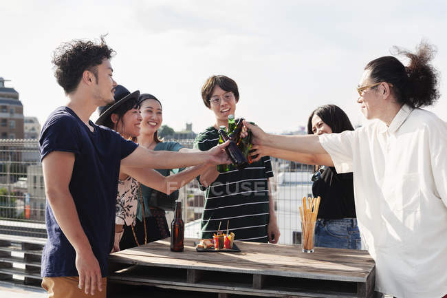 Group of young Japanese men and women standing on rooftop in urban setting, toasting beer bottles. — Stock Photo