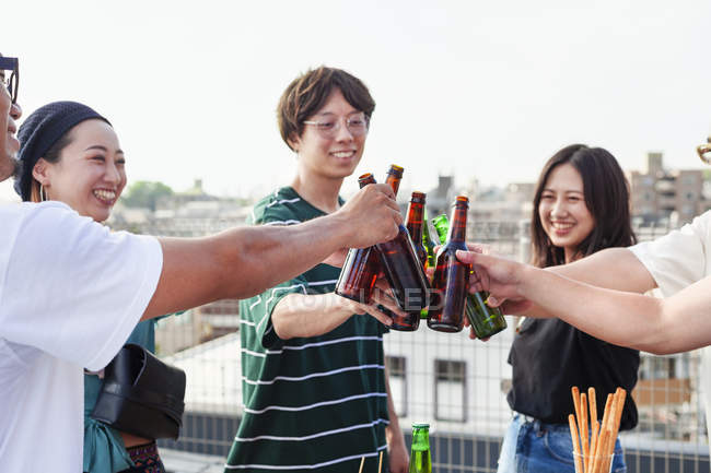 Group of young Japanese men and women standing on rooftop in urban setting, toasting beer bottles. — Stock Photo