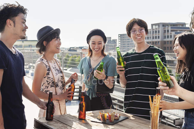 Gruppe junger japanischer Männer und Frauen steht auf einem Hausdach in urbaner Umgebung und trinkt Bier mit Snacks. — Stockfoto