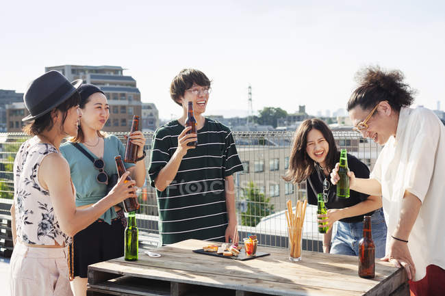 Group of young Japanese men and women standing on rooftop in urban setting, drinking beer with snacks. — Stock Photo