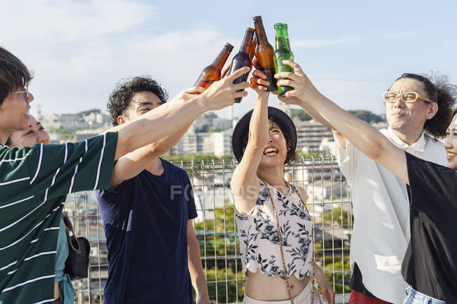 Gruppe junger japanischer Männer und Frauen steht auf einem Hausdach in urbaner Umgebung und prostet Bierflaschen zu. — Stockfoto