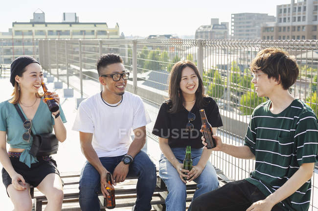Grupo de jovens homens e mulheres japoneses sentados no telhado em ambiente urbano, bebendo cerveja . — Fotografia de Stock