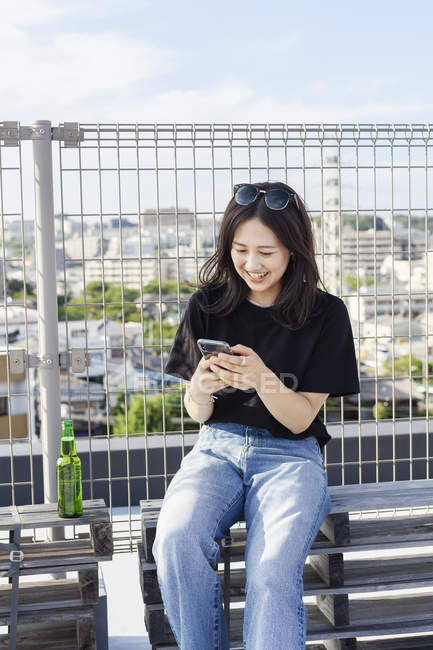 Young Japanese woman sitting on rooftop in urban setting and using mobile phone. — Stock Photo