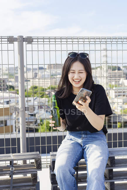 Молодая японка сидит на крыше в городской обстановке и пользуется мобильным телефоном . — стоковое фото