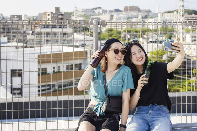 Two young Japanese women sitting on rooftop in urban setting, taking selfie with mobile phone and holding beer bottles. — Stock Photo