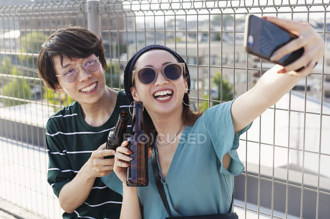 Young Japanese man and woman sitting on rooftop in urban setting, taking selfie with mobile phone. — Stock Photo