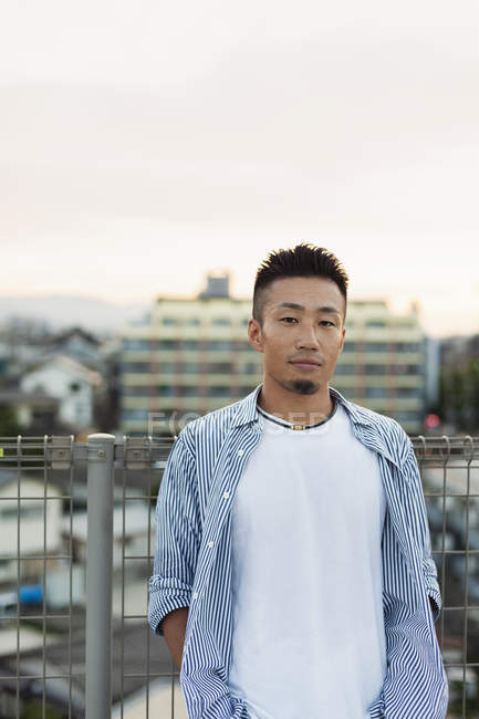 Junger Japaner steht auf einem Dach in urbaner Umgebung und blickt in die Kamera. — Stockfoto