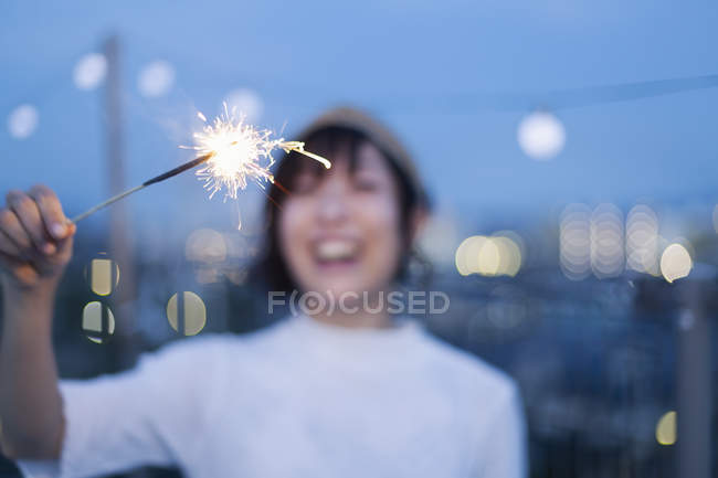 Sonriente joven japonesa sosteniendo chispeante en la azotea en el entorno urbano . - foto de stock