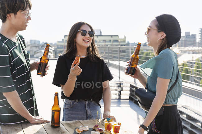 Jovem japonês homem e mulheres de pé no telhado em ambiente urbano, beber cerveja com lanches . — Fotografia de Stock