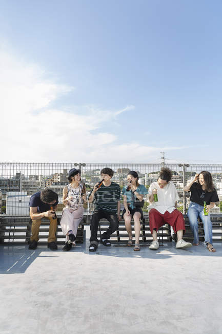 Gruppo sorridente di giovani giapponesi seduti con bottiglie di birra sul tetto in ambiente urbano . — Foto stock