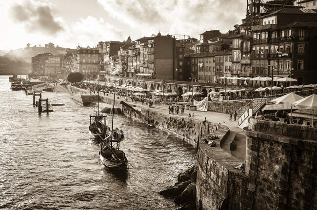 Port boats and freight barges moored beside a waterfront wall, and people on the promenade. Historic buildings. — Stock Photo
