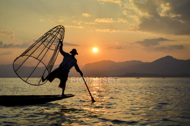 Fischer balanciert auf einem Bein auf einem Boot, hält einen großen Fischerkorb in der Hand und angelt auf traditionelle Weise auf dem Inle-See bei Sonnenuntergang, Myanmar. — Stockfoto