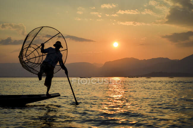 Традиционный рыбак балансирует на одной ноге на лодке, держа в руках рыболовную корзину, ловит рыбу на озере Инле на закате, Мьянма. — стоковое фото