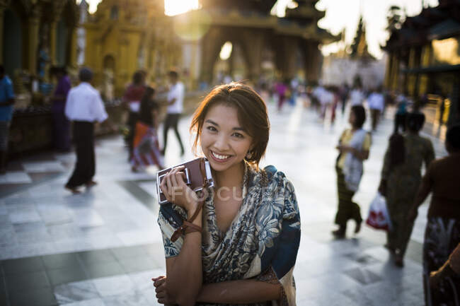 Молода жінка стоїть на міській площі, тримає стару камеру, посміхається на камеру . — стокове фото