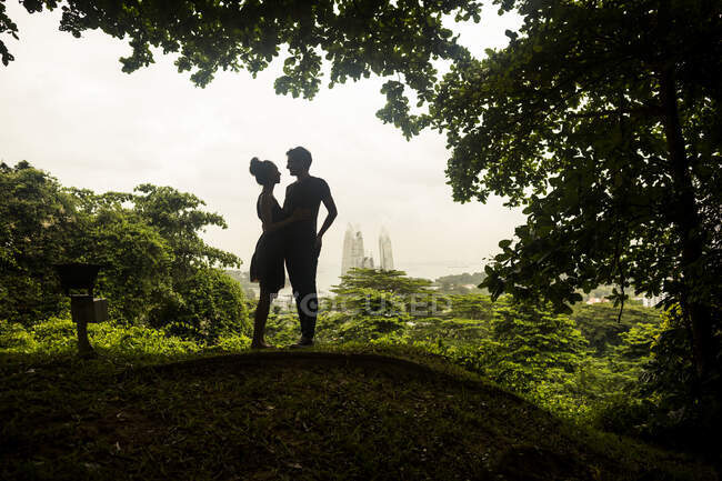 Silhueta de casal jovem de pé debaixo de árvores em uma floresta, arranha-céus à distância. — Fotografia de Stock