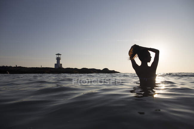 Rückansicht einer im Ozean badenden Frau bei Sonnenuntergang, Leuchtturm in der Ferne. — Stockfoto