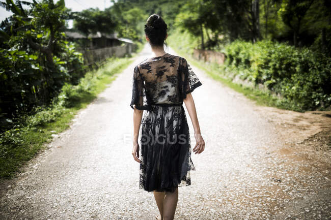 Rear view of woman wearing black lace dress walking down a rural country road. — Stock Photo